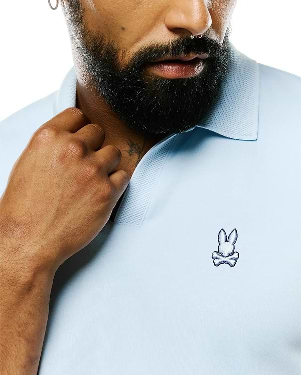 Mens Nasser Pima Johnny Polo in light blue Pima cotton jersey with a classic fit, featuring a textured pique collar and a small embroidered white and navy Psycho Bunny skull logo on the left chest.