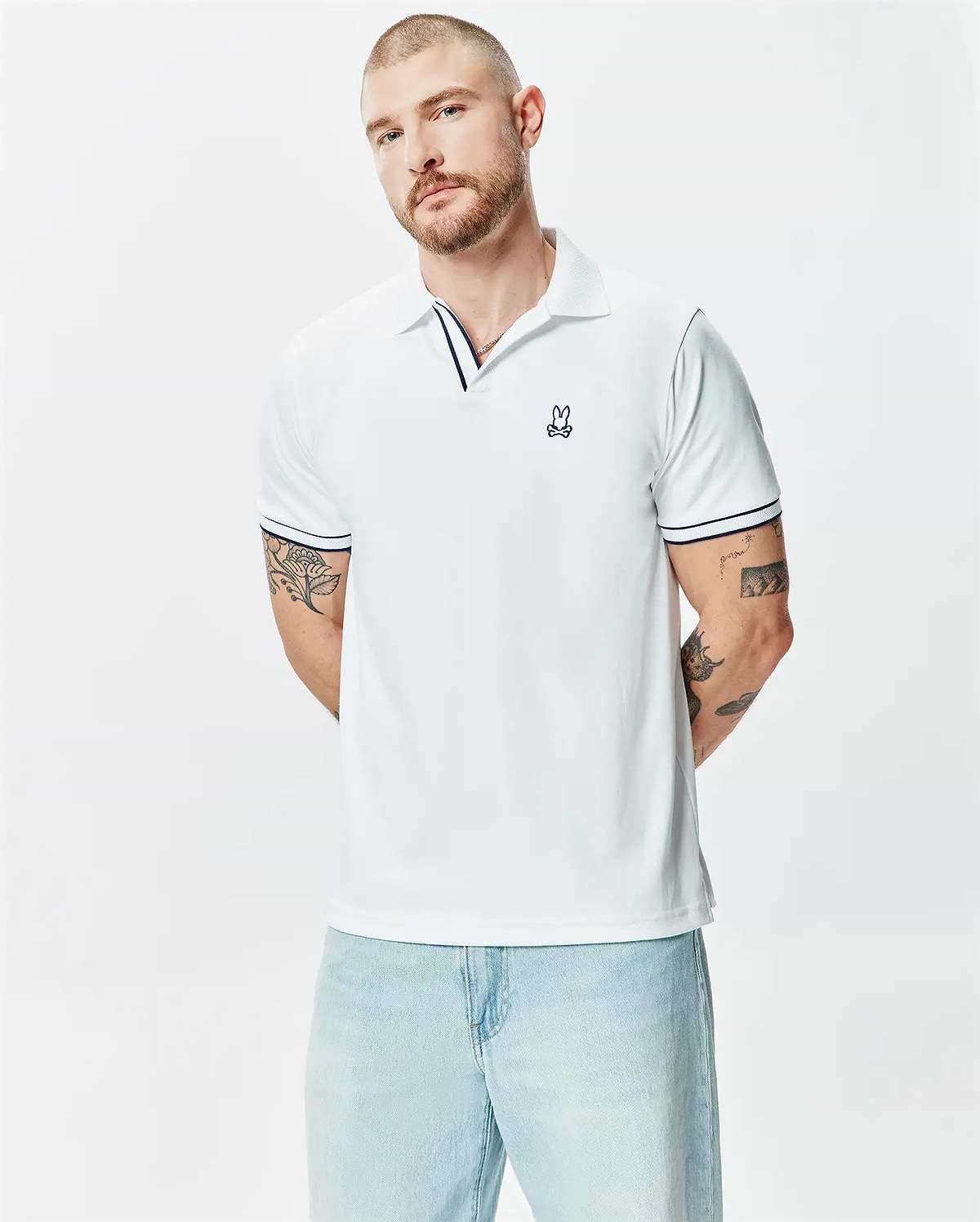 Mens Nasser Polo, white pique knit, classic fit, with navy blue striped collar and cuffs, featuring a small embroidered Psycho Bunny skull logo on the left chest.