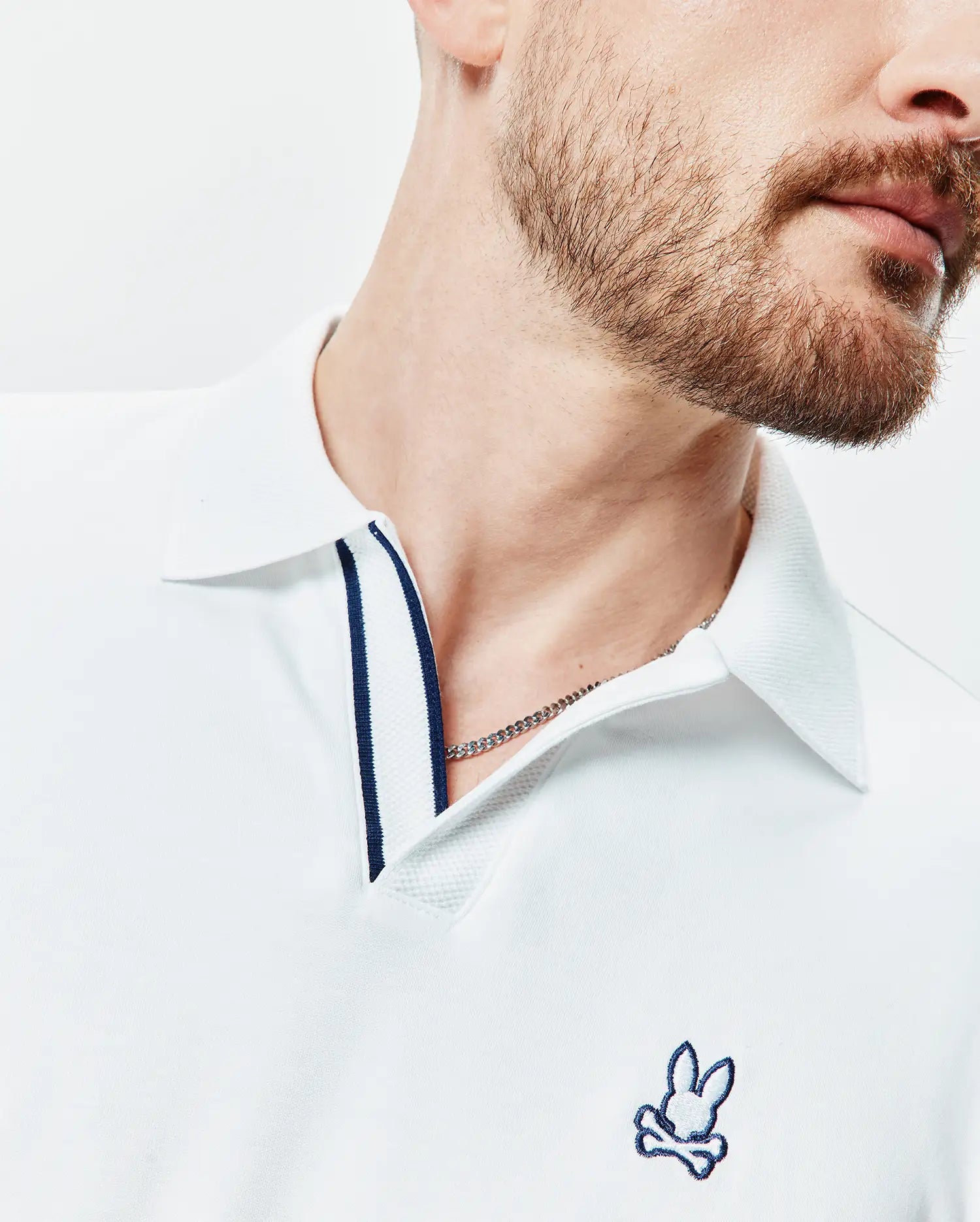 White Mens Nasser Polo, a classic fit pique knit shirt with a navy blue striped placket and a small embroidered blue and white Psycho Bunny skull logo on the left chest.