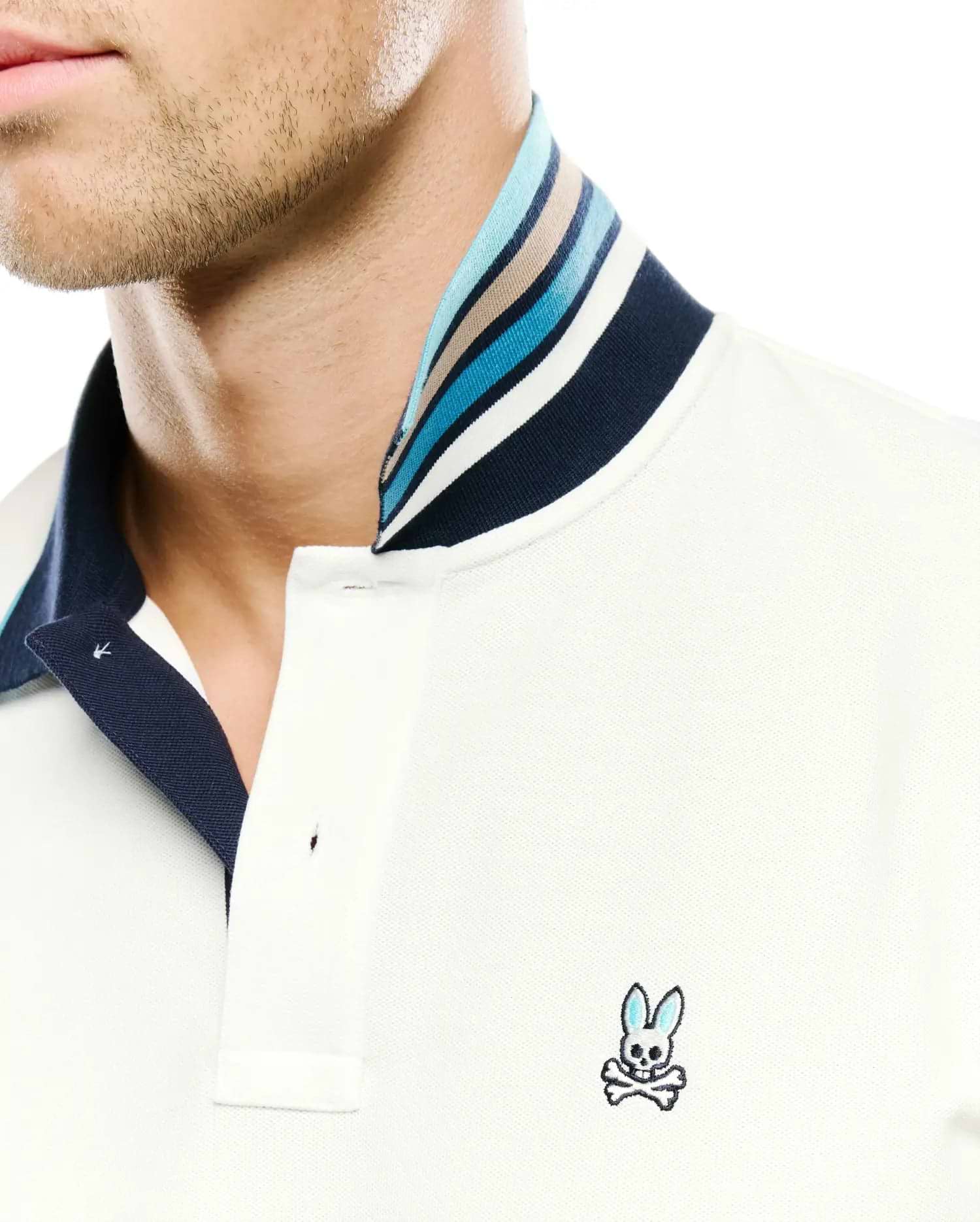 Men's Klaus Pique Polo in cream, featuring a classic fit, two-button placket, and a navy blue collar with multi-colored stripes in blues, white, and tan. The small, embroidered Psycho Bunny skull logo with blue ears is on the left chest. (2)