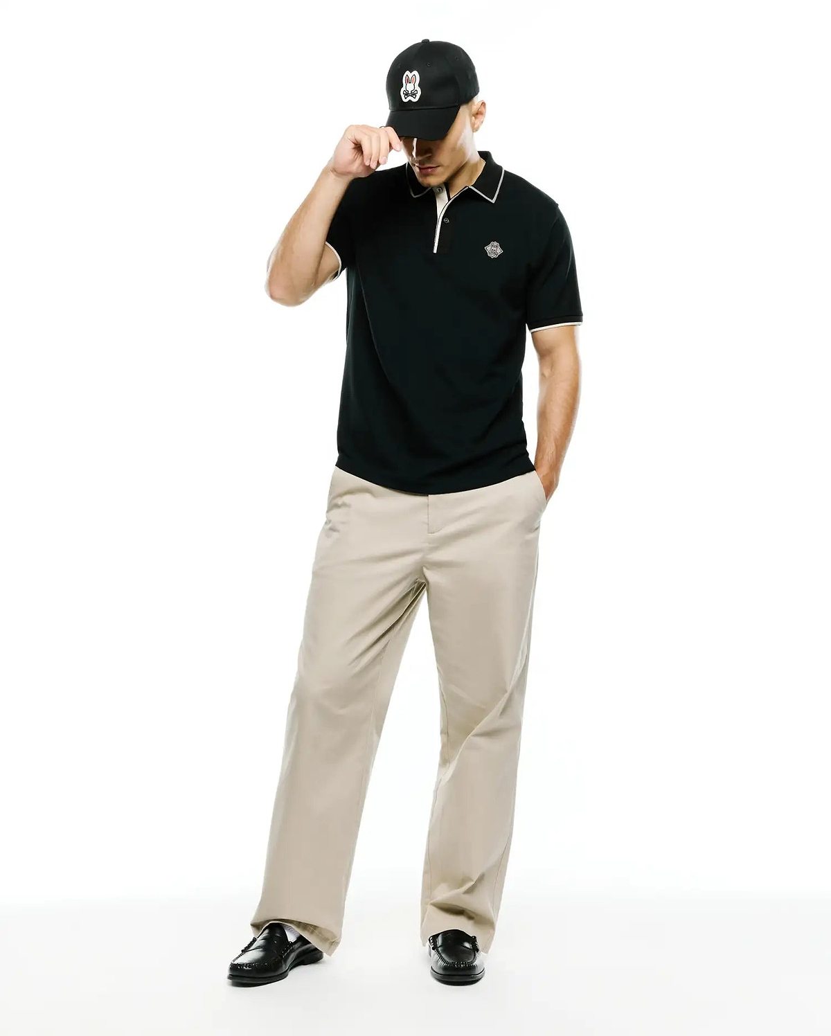 Mens Robin Pima Piqué Polo in classic black with white collar and cuff tipping, featuring a small embroidered Psycho Bunny skull logo on the left chest, paired with khaki trousers and a black Psycho Bunny cap.