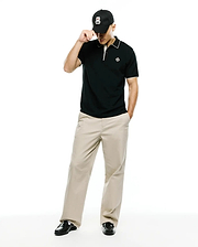 Mens Robin Pima Piqué Polo in classic black with white collar and cuff tipping, featuring a small embroidered Psycho Bunny skull logo on the left chest, paired with khaki trousers and a black Psycho Bunny cap.