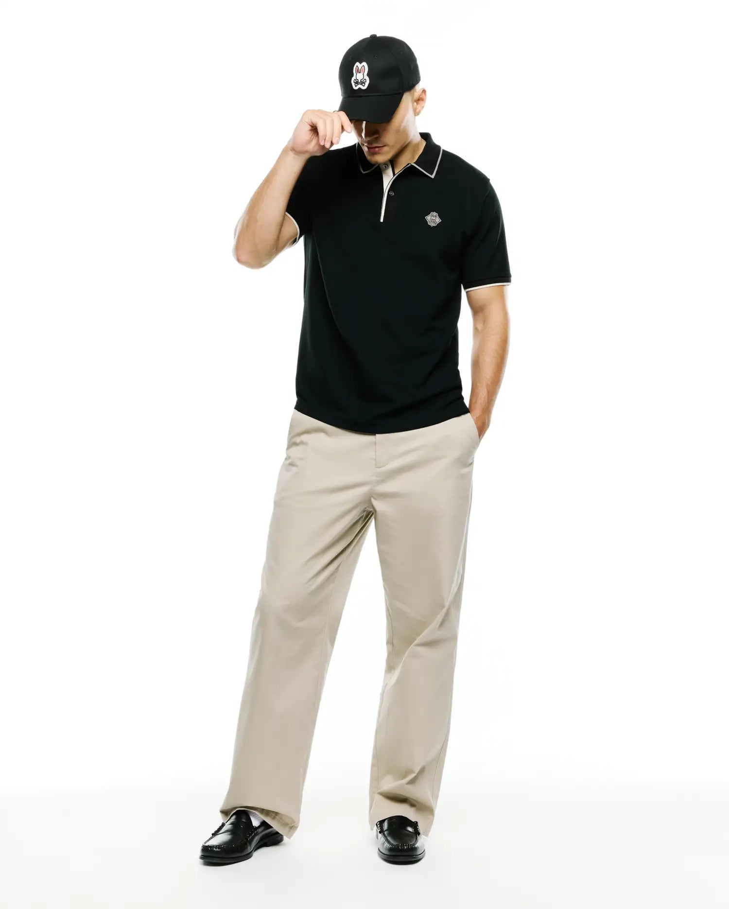 Mens Robin Pima Piqué Polo in classic black with white collar and cuff tipping, featuring a small embroidered Psycho Bunny skull logo on the left chest, paired with khaki trousers and a black Psycho Bunny cap.