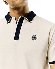 Mens Robin Pique Polo in cream, featuring a classic fit, pique knit texture, navy contrast collar, two-button placket with navy trim, ribbed navy sleeve cuffs, and a small embroidered Psycho Bunny skull logo on the left chest.