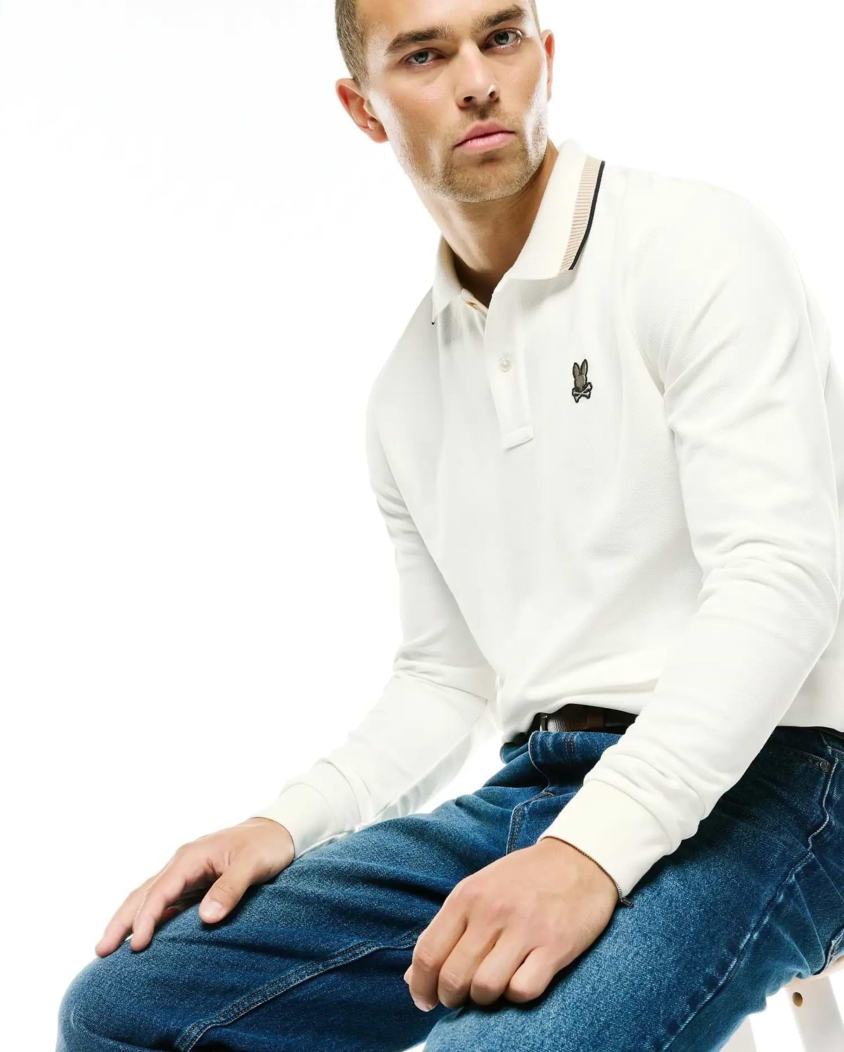 Men's Grover Pique Longsleeve Polo in white, featuring a classic fit, textured pique fabric, and a small embroidered Psycho Bunny skull logo on the left chest, with a striped collar in black and tan. (3)