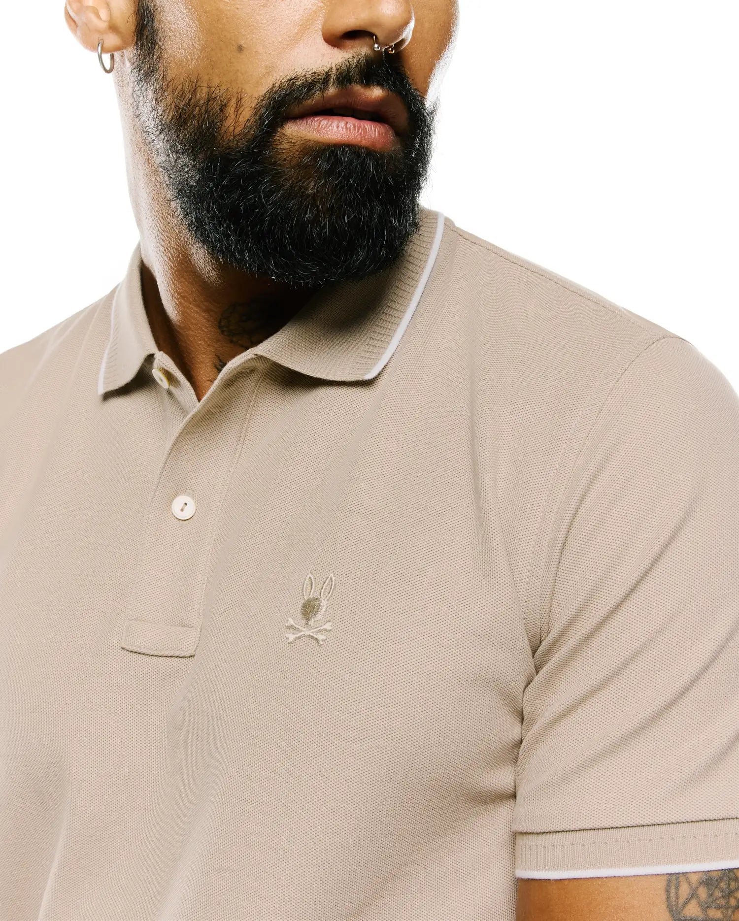 Mens Ringston Pique Polo in classic fit beige pique fabric, featuring a two-button placket, white tipping on the ribbed collar and cuffs, and a small embroidered Psycho Bunny skull logo on the left chest.