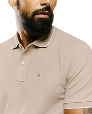 Mens Ringston Pique Polo in classic fit beige pique fabric, featuring a two-button placket, white tipping on the ribbed collar and cuffs, and a small embroidered Psycho Bunny skull logo on the left chest.