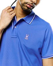 Mens Atley Pique Polo in royal blue, featuring a classic fit, pique knit texture, white and purple striped collar and cuffs, and a small embroidered purple Psycho Bunny skull logo on the left chest. (2)