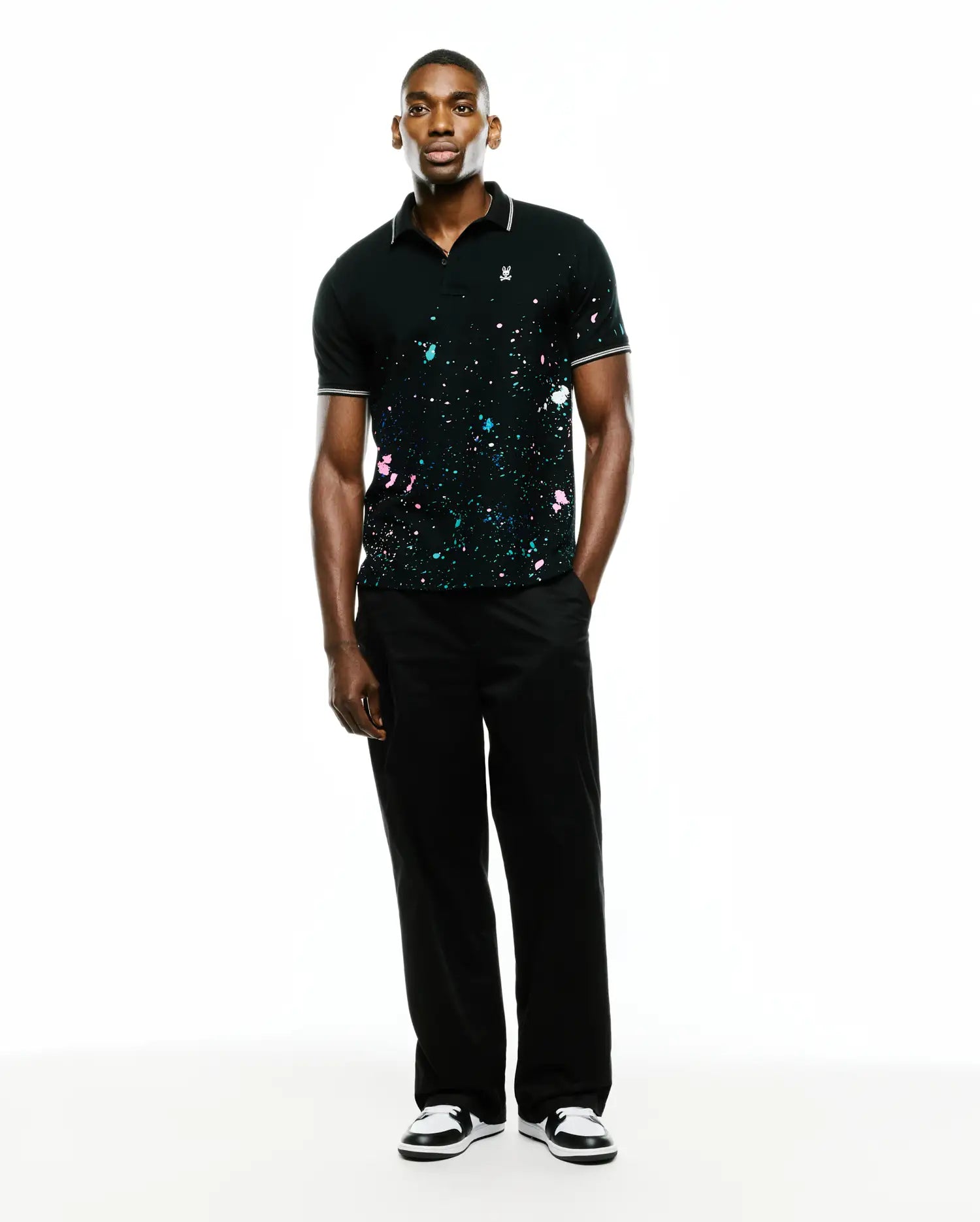 Mens Malcolm Splatter Polo, a black pique polo with classic fit, featuring vibrant pink, blue, white paint splatter, white-tipped ribbed collar/cuffs, and small embroidered Psycho Bunny skull logo on left chest. (4)