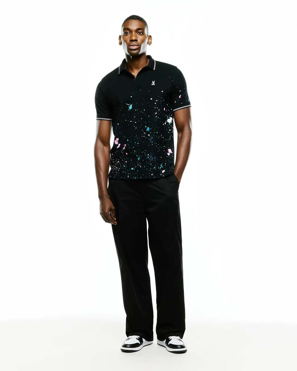 Mens Malcolm Splatter Polo, a black pique polo with classic fit, featuring vibrant pink, blue, white paint splatter, white-tipped ribbed collar/cuffs, and small embroidered Psycho Bunny skull logo on left chest. (4)