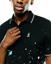 Mens Malcolm Splatter Polo, black pique knit polo with pink, turquoise, white splatter paint, small white embroidered Psycho Bunny skull logo on chest, and white striped collar and cuffs. (2)