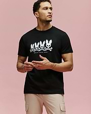 A man with light skin and short curly hair wears beige pants and the Psycho Bunny MENS ARCHIVE GRADIANT BUNNY GRAPHIC TEE (MT0200029), a black Pima cotton shirt with four white Bunny skull graphics and "BOLD SINCE 2005" on a pink background.