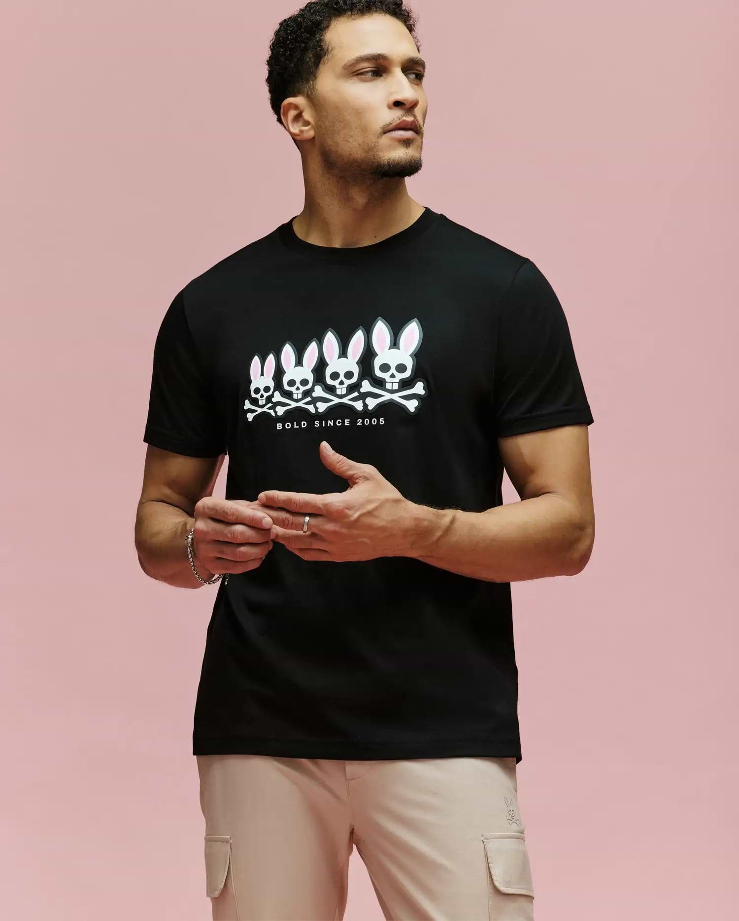 A man with light skin and short curly hair wears beige pants and the Psycho Bunny MENS ARCHIVE GRADIANT BUNNY GRAPHIC TEE (MT0200029), a black Pima cotton shirt with four white Bunny skull graphics and "BOLD SINCE 2005" on a pink background.