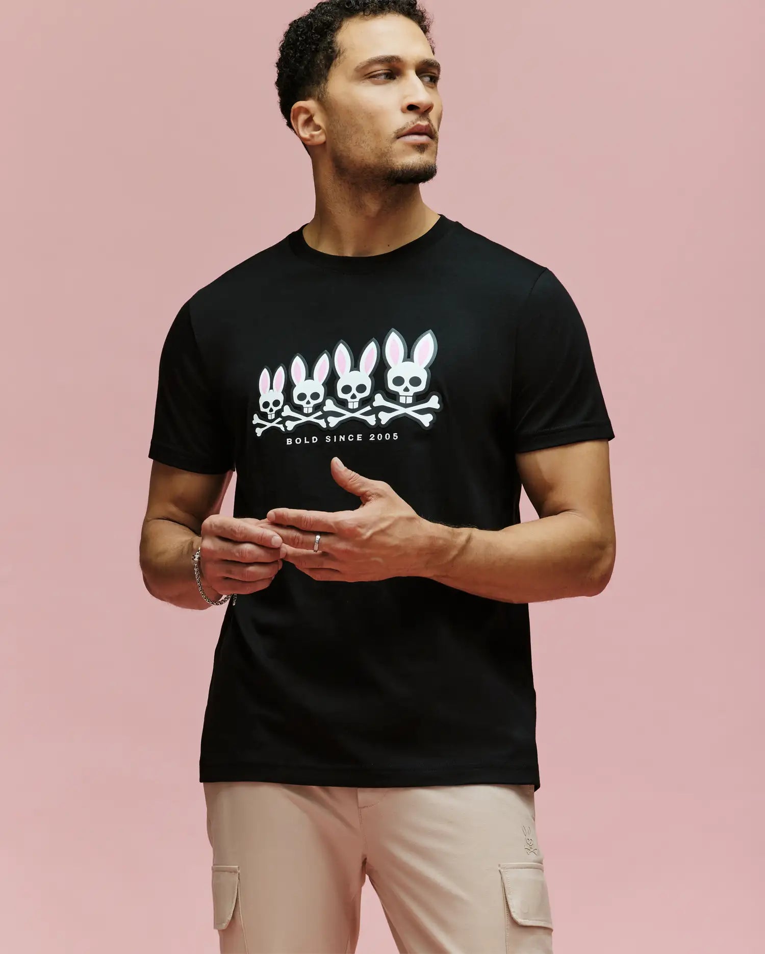 A man with light skin and short curly hair wears beige pants and the Psycho Bunny MENS ARCHIVE GRADIANT BUNNY GRAPHIC TEE (MT0200029), a black Pima cotton shirt with four white Bunny skull graphics and "BOLD SINCE 2005" on a pink background.