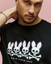 A man poses against a pink background, wearing the Psycho Bunny MENS ARCHIVE GRADIANT BUNNY GRAPHIC TEE (MT0200029), a Pima cotton shirt featuring four Bunny skull graphics and "BOLD SINCE 2005" text below the design.