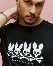 A man poses against a pink background, wearing the Psycho Bunny MENS ARCHIVE GRADIANT BUNNY GRAPHIC TEE (MT0200029), a Pima cotton shirt featuring four Bunny skull graphics and "BOLD SINCE 2005" text below the design.