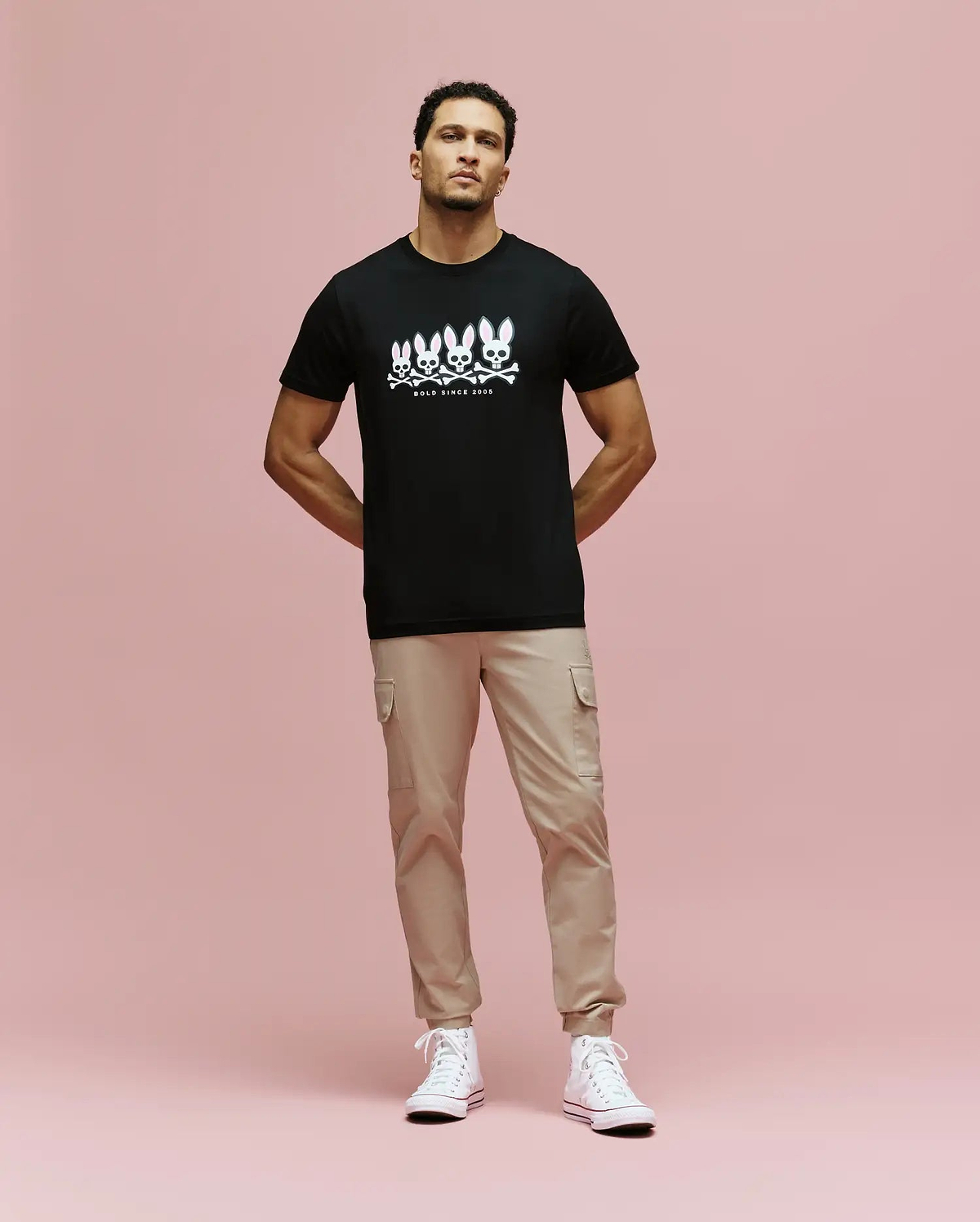 A man with short hair stands against a pink background in a Psycho Bunny MENS ARCHIVE GRADIENT BUNNY GRAPHIC TEE (MT0200029), beige cargo pants, and white high-top sneakers. He has a neutral expression and his hands are in his pockets.