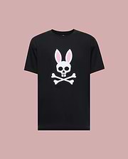 The Psycho Bunny MENS ARCHIVE BUNNY GRAPHIC TEE (MT0200031) is a black 100% Pima cotton shirt made in Peru, featuring a bold white skull with rabbit ears and pink inner accents, displayed against a light mauve background.