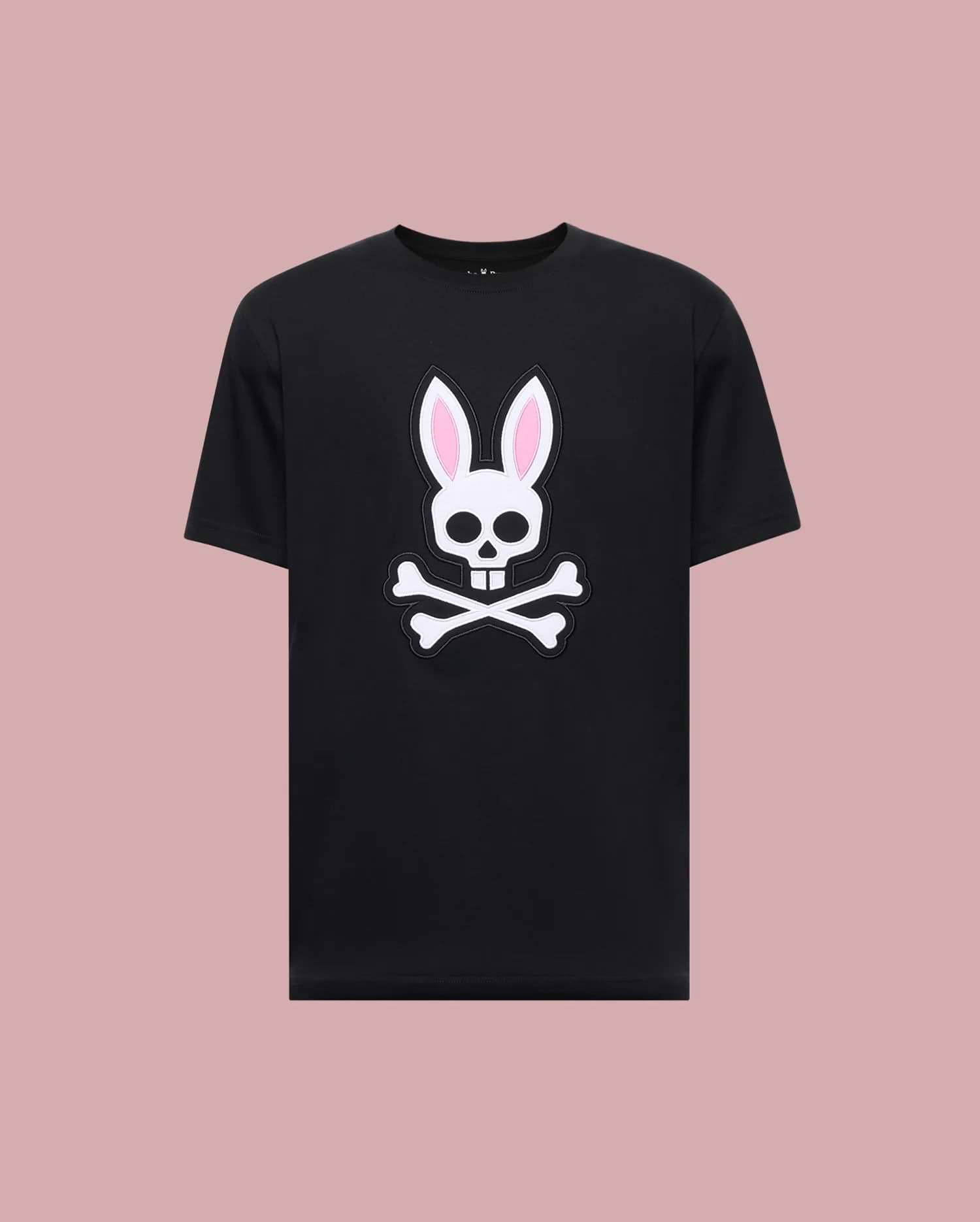 The Psycho Bunny MENS ARCHIVE BUNNY GRAPHIC TEE (MT0200031) is a black 100% Pima cotton shirt made in Peru, featuring a bold white skull with rabbit ears and pink inner accents, displayed against a light mauve background.