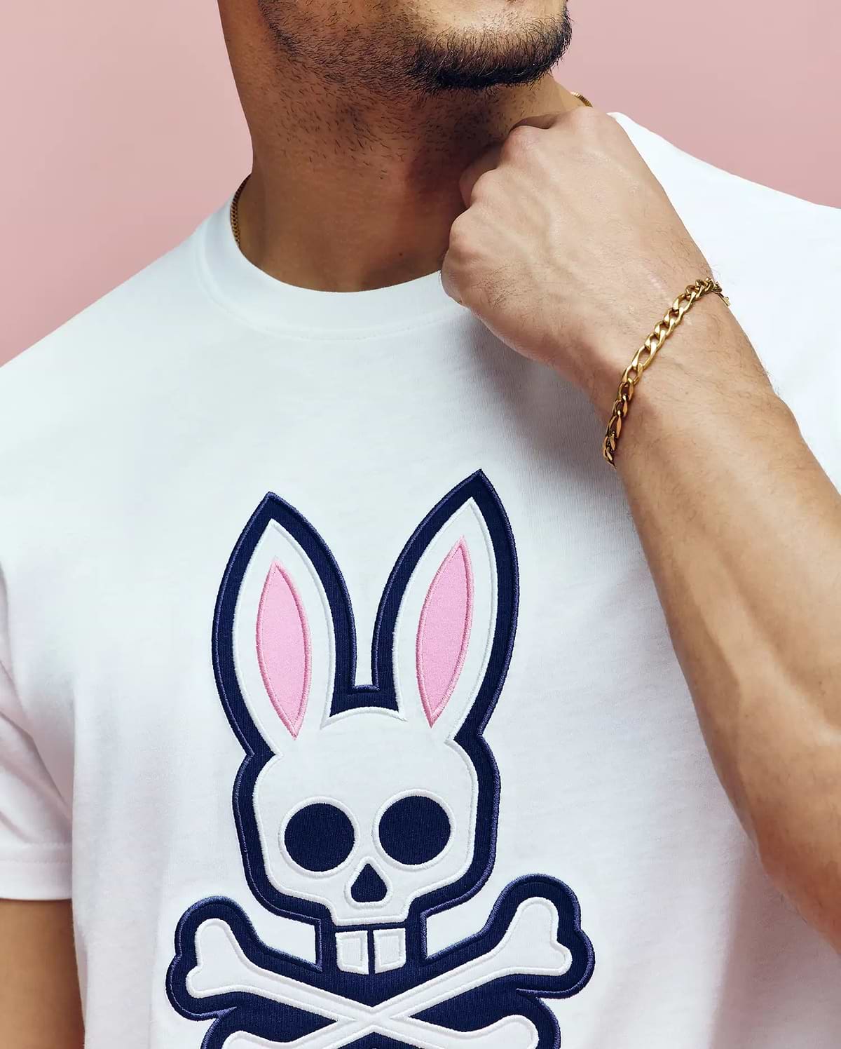 A man wears the Psycho Bunny MENS ARCHIVE BUNNY GRAPHIC TEE (MT0200031), a white Pima cotton shirt with a large skull and crossbones bunny graphic, gold chain bracelet, touching his neck against a pink background.