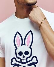 A man wears the Psycho Bunny MENS ARCHIVE BUNNY GRAPHIC TEE (MT0200031), a white Pima cotton shirt with a large skull and crossbones bunny graphic, gold chain bracelet, touching his neck against a pink background.