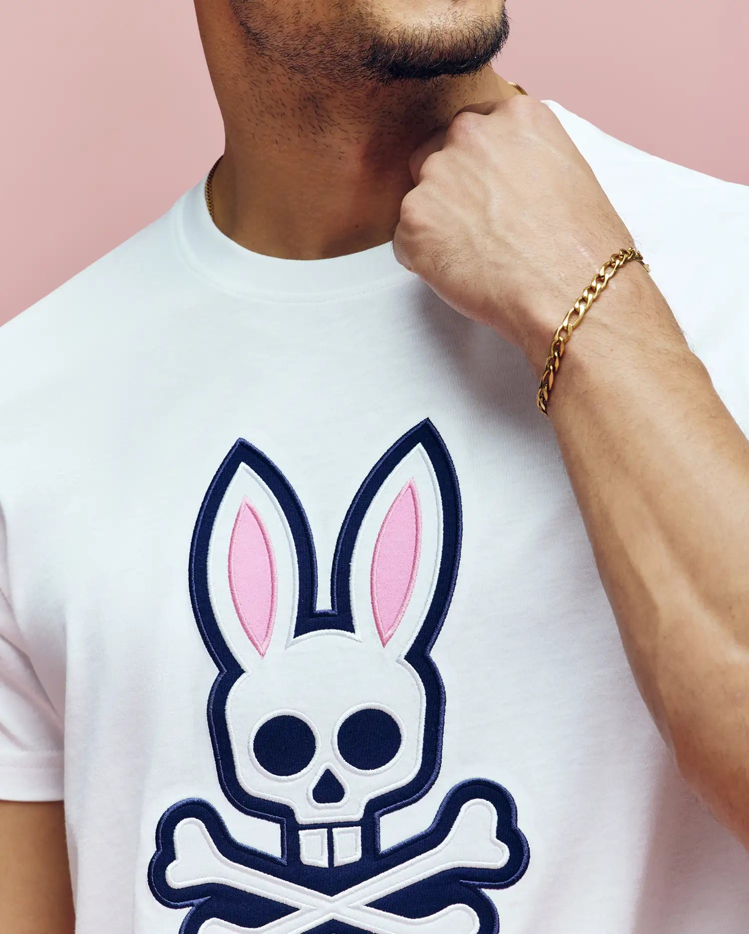 A man wears the Psycho Bunny MENS ARCHIVE BUNNY GRAPHIC TEE (MT0200031), a white Pima cotton shirt with a large skull and crossbones bunny graphic, gold chain bracelet, touching his neck against a pink background.