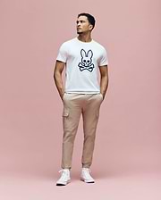 A man stands against a pink background, wearing the Psycho Bunny MENS ARCHIVE BUNNY GRAPHIC TEE (MT0200031), beige cargo pants, and white sneakers. His hands are in his pockets as he looks slightly to the side.