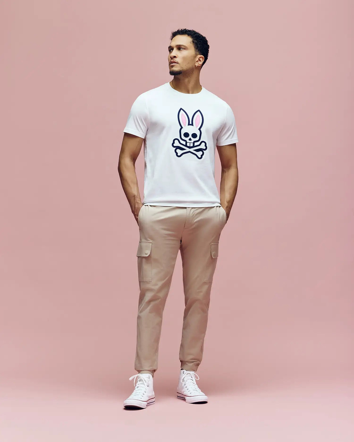 A man stands against a pink background, wearing the Psycho Bunny MENS ARCHIVE BUNNY GRAPHIC TEE (MT0200031), beige cargo pants, and white sneakers. His hands are in his pockets as he looks slightly to the side.