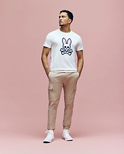 A man stands against a pink background, wearing the Psycho Bunny MENS ARCHIVE BUNNY GRAPHIC TEE (MT0200031), beige cargo pants, and white sneakers. His hands are in his pockets as he looks slightly to the side.