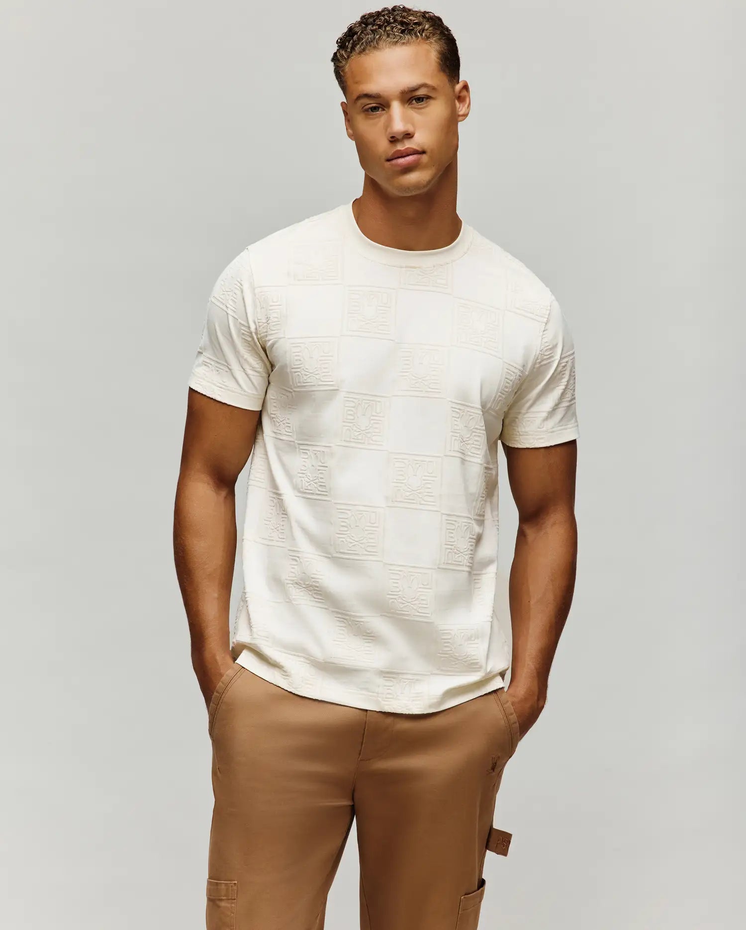 Mens Gio Tee, a cream classic fit jersey t-shirt featuring an all-over embossed checkerboard pattern with the iconic Psycho Bunny skull logo in each square.