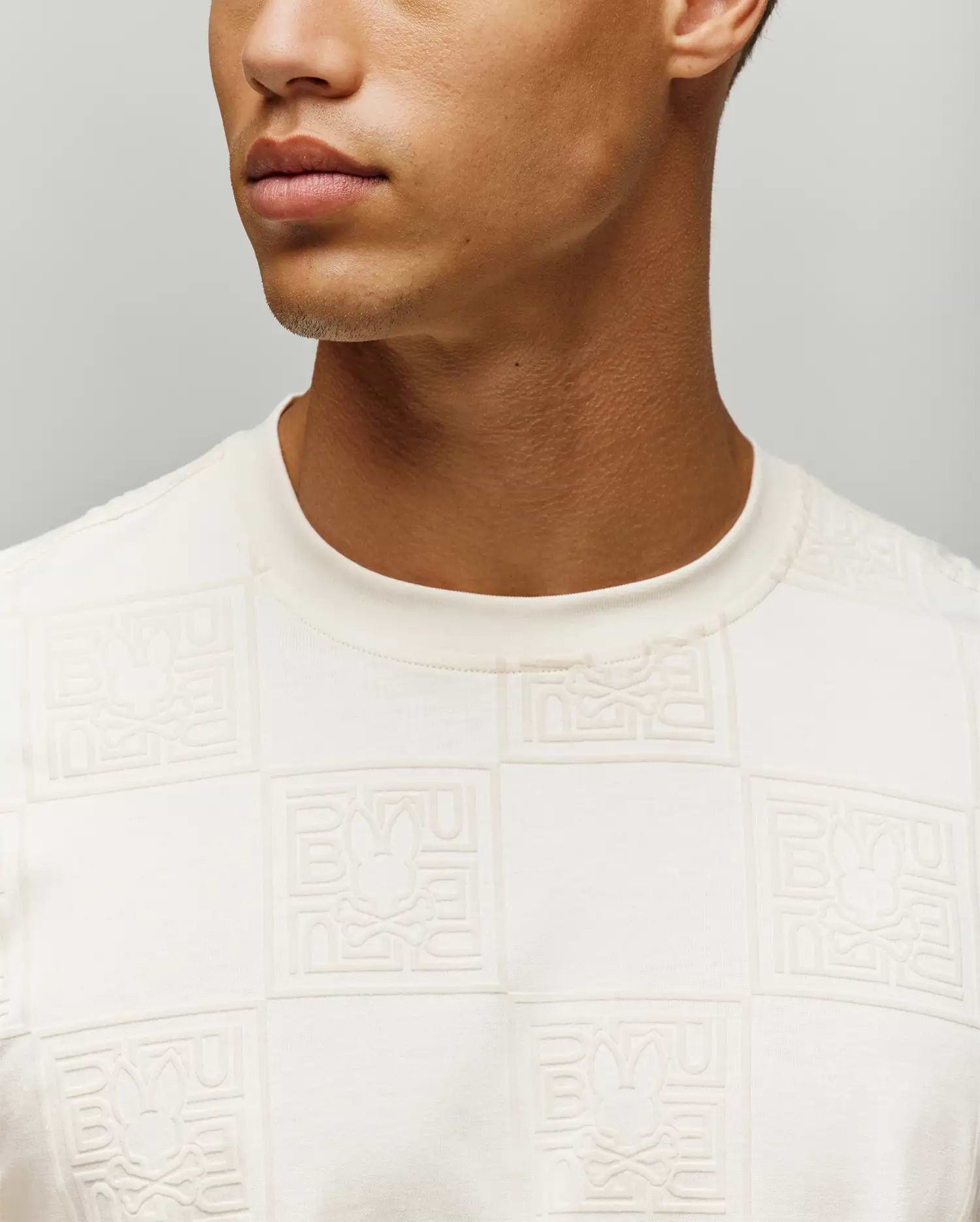 Mens Gio Tee in cream, featuring a classic fit and soft jersey fabric with an all-over embossed checkerboard pattern of the Psycho Bunny skull logo and brand name.
