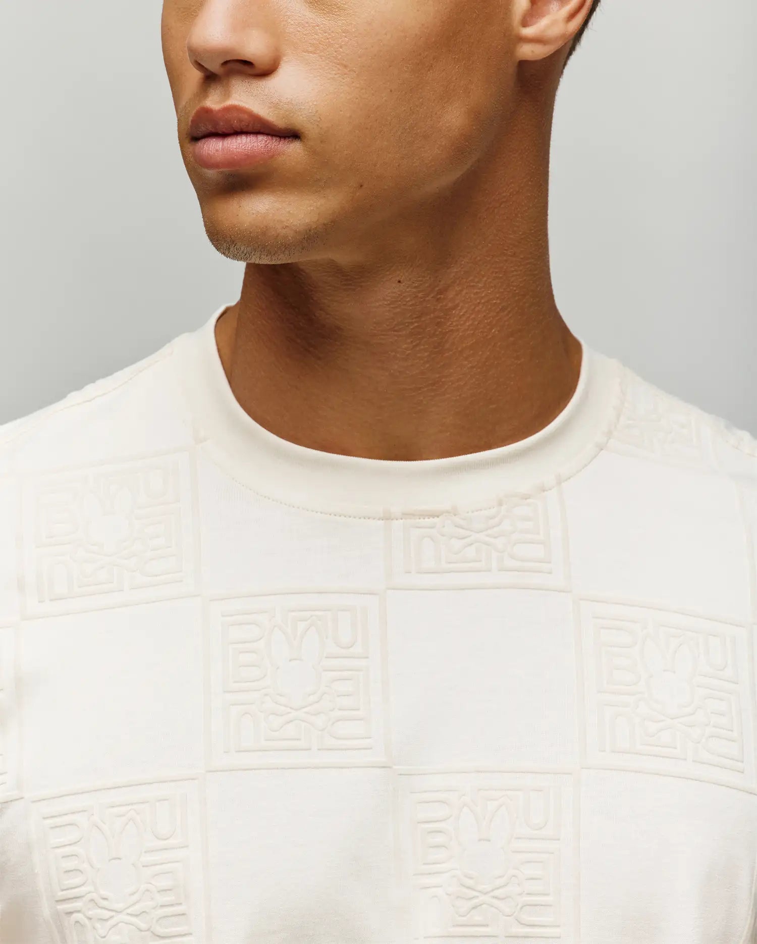 Mens Gio Tee in cream, featuring a classic fit and soft jersey fabric with an all-over embossed checkerboard pattern of the Psycho Bunny skull logo and brand name.