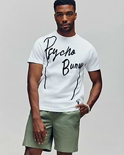A man wears a white Psycho Bunny MENS SOREN GRAPHIC TEE (MT0200180), made of 100% cotton with a flocked "Psycho Bunny" wordmark, and light green shorts, standing against a plain light gray background. (2)