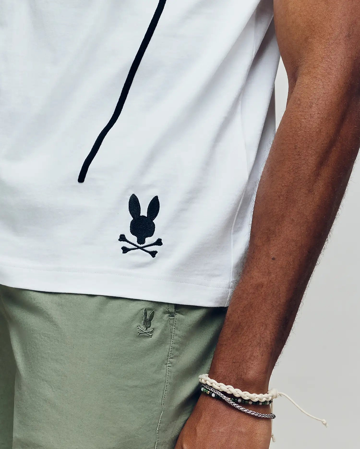 Wearing the Psycho Bunny MENS SOREN GRAPHIC TEE (MT0200180), a 100% cotton white tee with a black bunny and crossbones logo near the hem, and light green shorts, they rest their bracelet-adorned arm along the shirt’s side seam. (3)