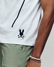 Wearing the Psycho Bunny MENS SOREN GRAPHIC TEE (MT0200180), a 100% cotton white tee with a black bunny and crossbones logo near the hem, and light green shorts, they rest their bracelet-adorned arm along the shirt’s side seam. (3)