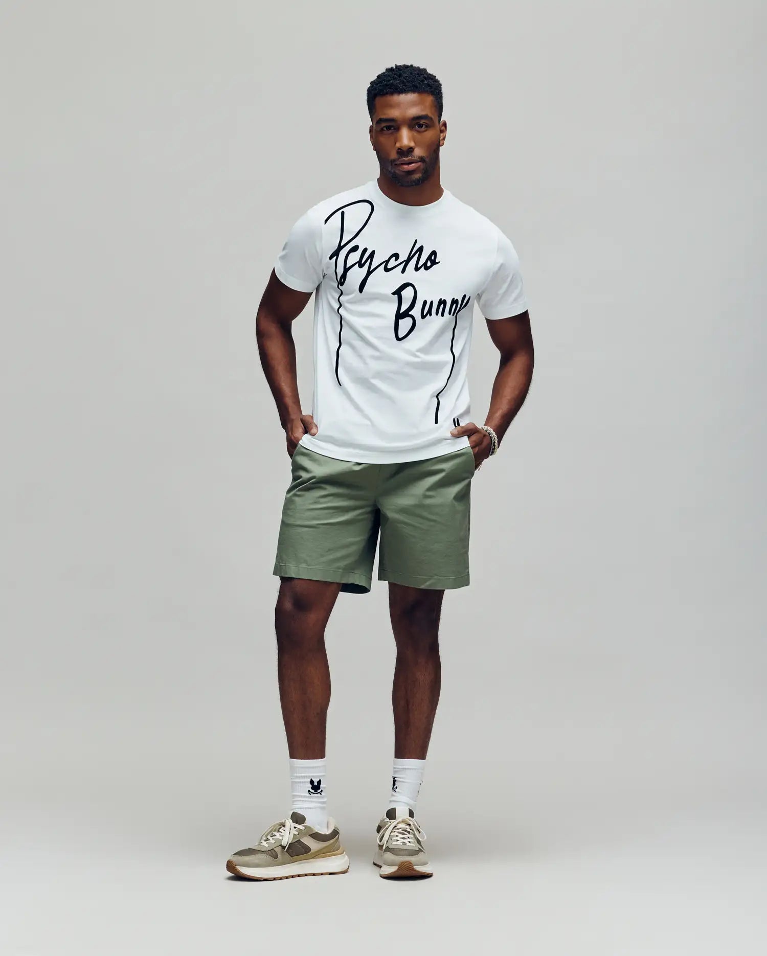 A man stands against a plain background wearing the Psycho Bunny MENS SOREN GRAPHIC TEE (MT0200180) in white, paired with light green shorts, white socks, and beige sneakers. His hands are in his pockets as he looks straight ahead. (4)