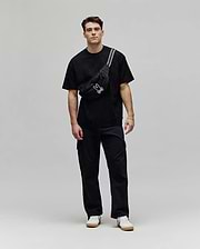 Mens Dustin Oversized Tee in black with a subtle textured checkerboard pattern, paired with a black crossbody fanny pack featuring a prominent white embroidered Psycho Bunny skull logo and white stripe detail.