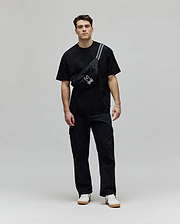 Mens Dustin Oversized Tee in black with a subtle textured checkerboard pattern, paired with a black crossbody fanny pack featuring a prominent white embroidered Psycho Bunny skull logo and white stripe detail.