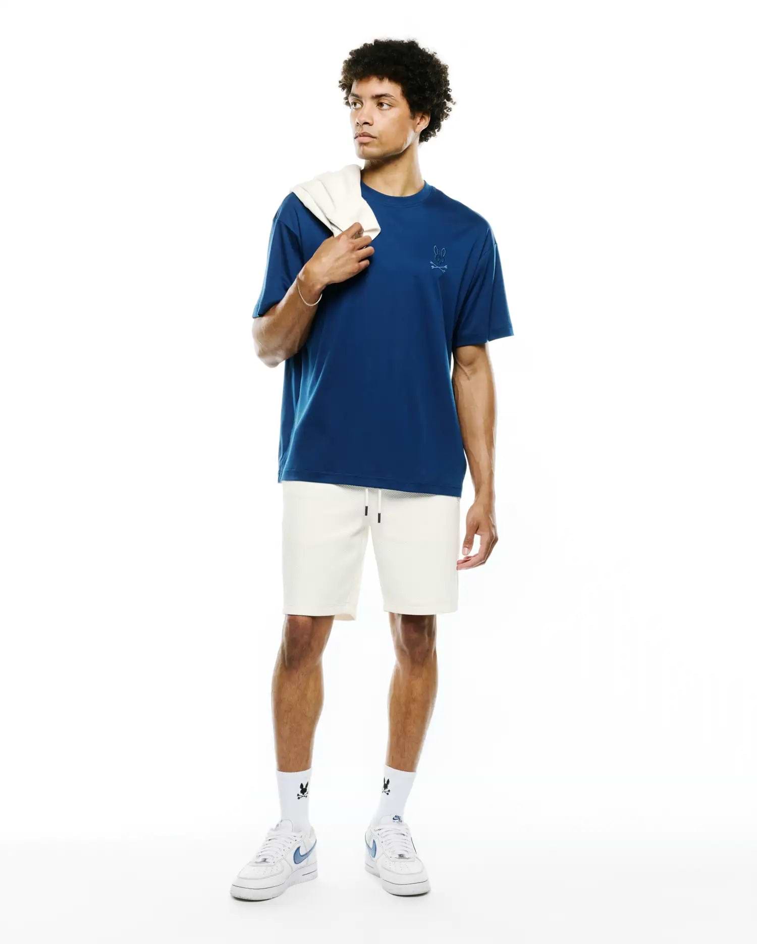 Mens Oscar Relaxed Fit Tee in deep blue jersey with a tonal embroidered Psycho Bunny skull logo on the left chest, paired with cream waffle knit shorts and white socks featuring black Psycho Bunny logos.