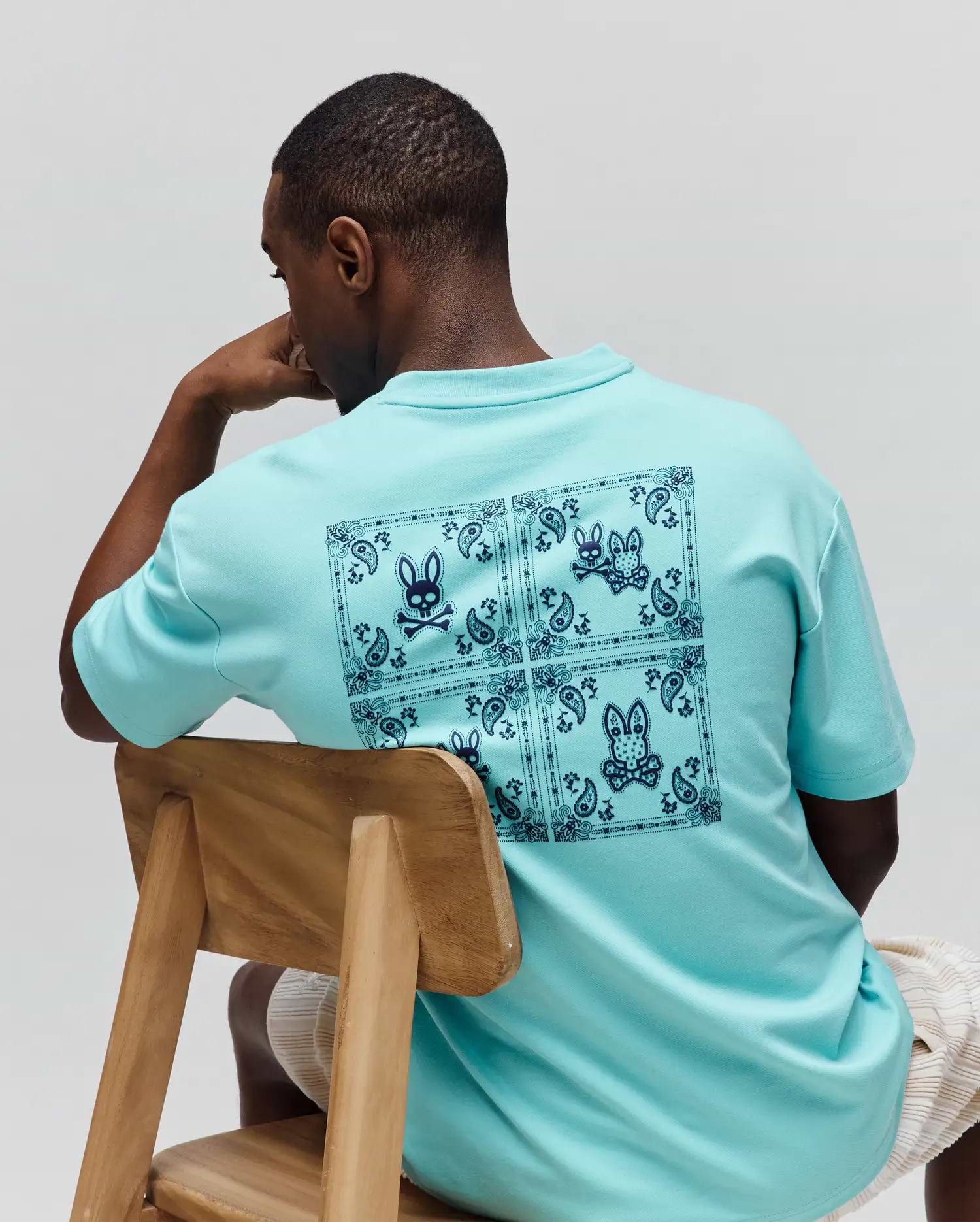Mens Luke Back Graphic Tee in aqua, featuring a relaxed fit and smooth jersey knit. A large dark blue bandana-style graphic with multiple Psycho Bunny skull logos covers the entire back.