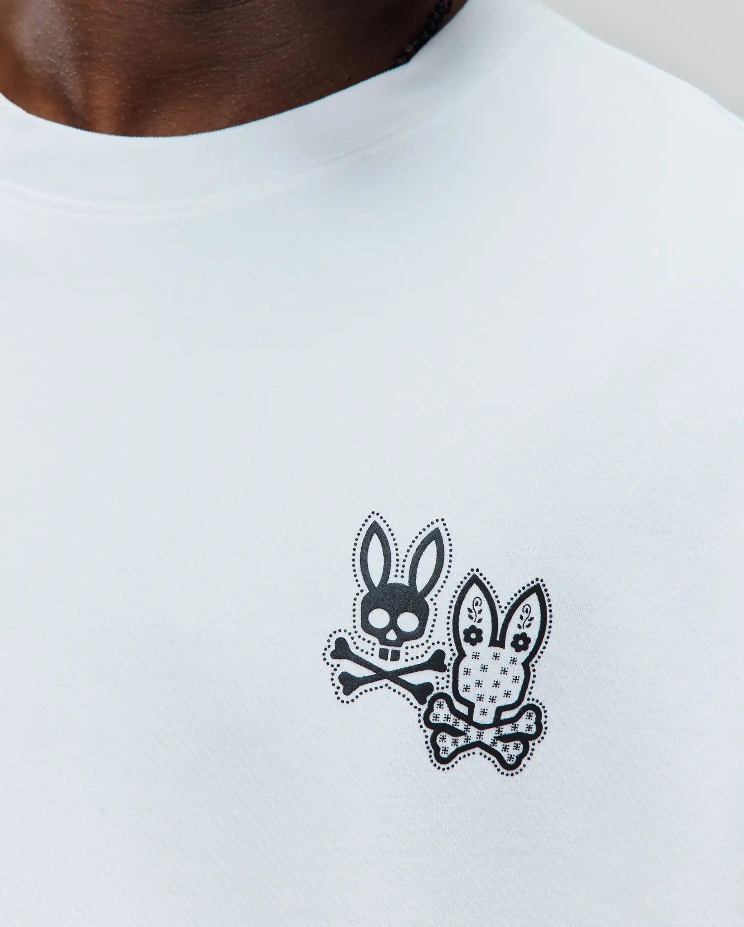 White jersey knit T-shirt with a small black graphic on the left chest, featuring two stylized Psycho Bunny skulls with dotted outlines, one classic with crossbones, the other with floral patterns. Mens Luke Back Graphic Tee.
