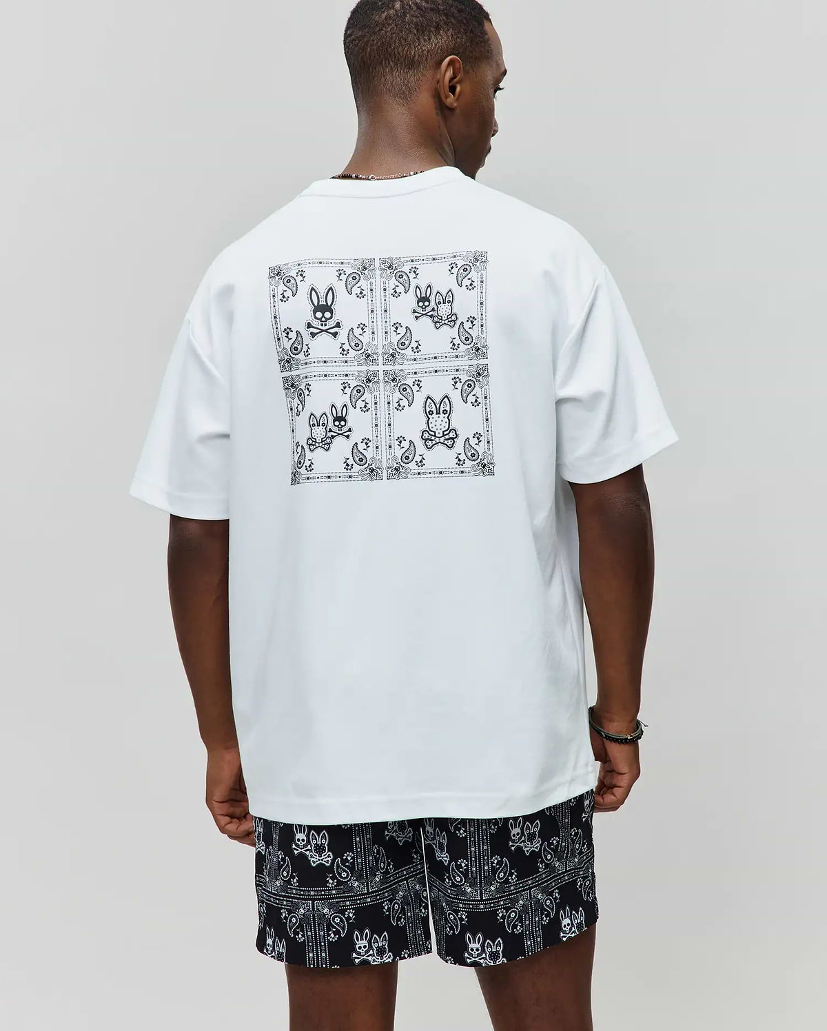 White Mens Luke Back Graphic Tee, relaxed jersey tee with a large black bandana graphic and four Psycho Bunny skull logos on the back, paired with black relaxed shorts with all-over white bandana-bunny skull pattern.