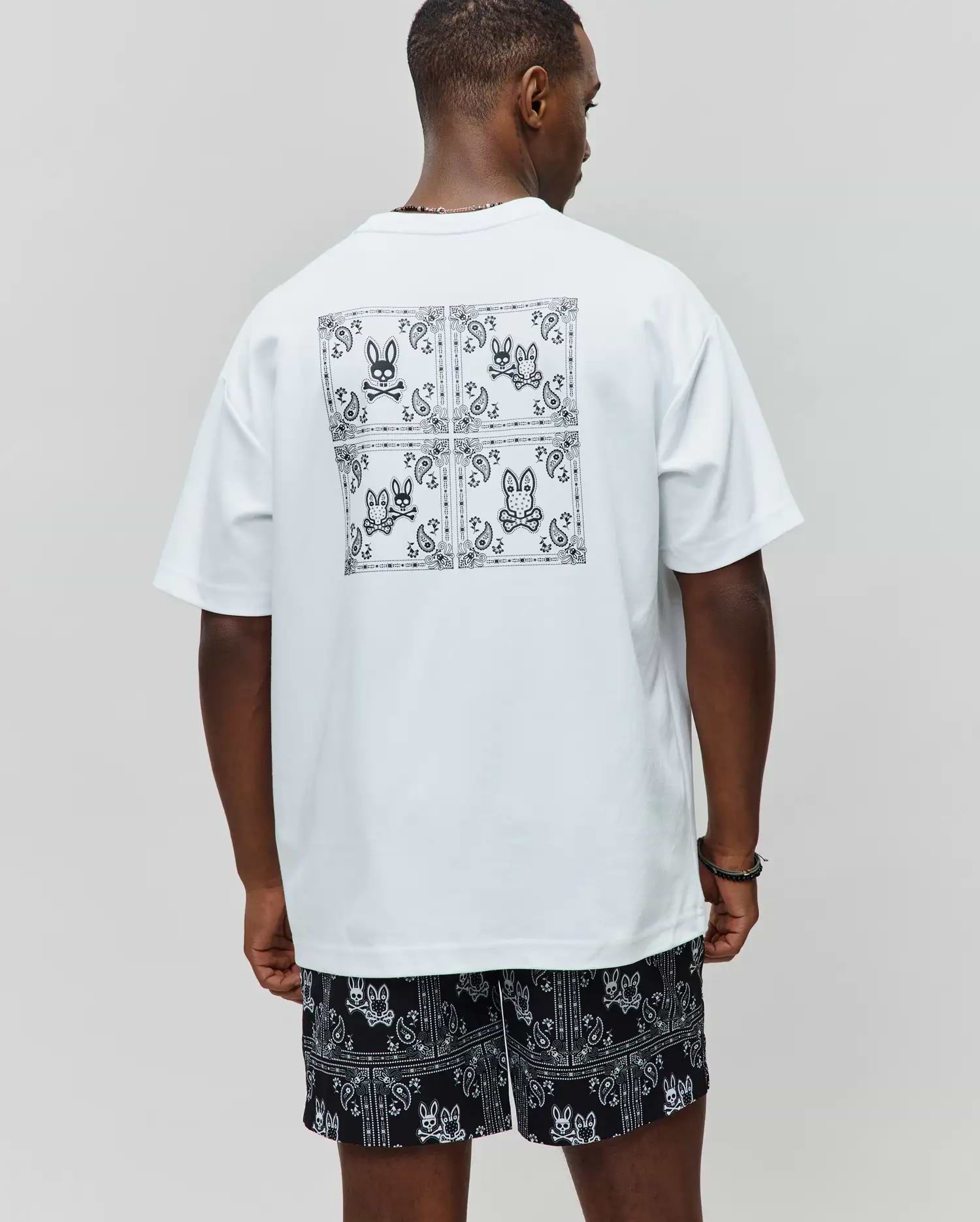 White Mens Luke Back Graphic Tee, relaxed jersey tee with a large black bandana graphic and four Psycho Bunny skull logos on the back, paired with black relaxed shorts with all-over white bandana-bunny skull pattern.