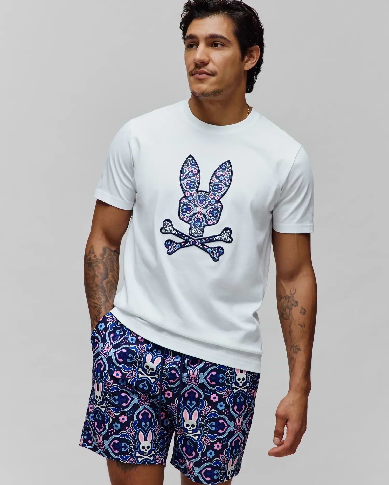 White Mens Luke Graphic Tee, classic fit, with a large ornate blue, pink, and purple floral Psycho Bunny skull chest graphic. Navy shorts feature an all-over matching bunny skull pattern.