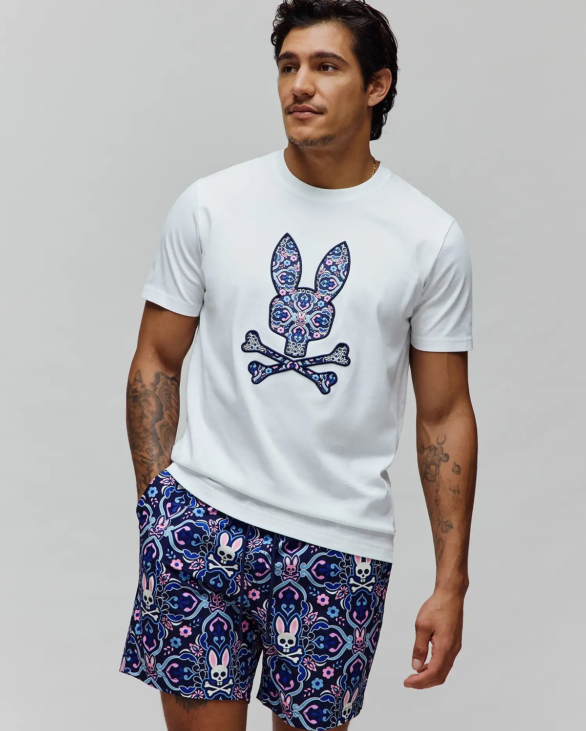 White Mens Luke Graphic Tee, classic fit, with a large ornate blue, pink, and purple floral Psycho Bunny skull chest graphic. Navy shorts feature an all-over matching bunny skull pattern.
