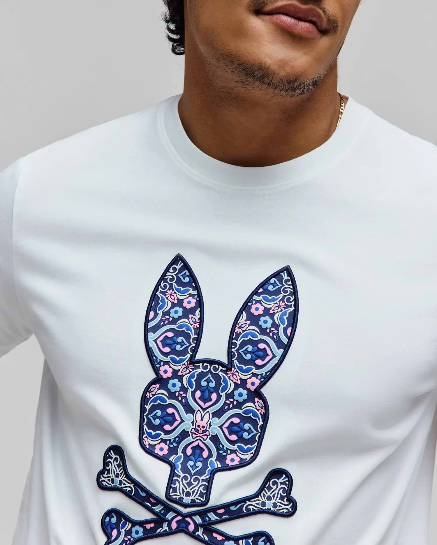 Mens Luke Graphic Tee, white jersey t-shirt with a large raised chest graphic of the iconic Psycho Bunny skull logo, filled with a dark blue, light blue, and pink floral paisley pattern.