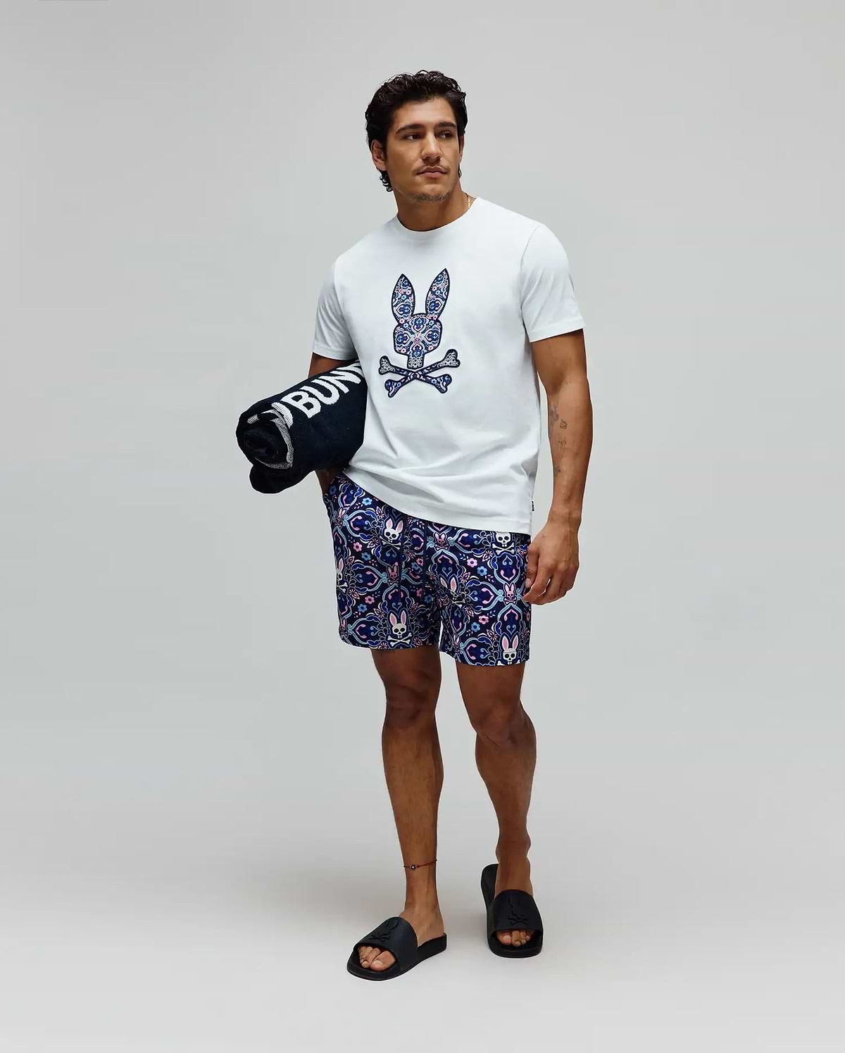 Mens Luke Graphic Tee, a classic fit white jersey t-shirt with a large, vibrant patterned Psycho Bunny skull chest graphic, paired with relaxed navy shorts featuring an all-over bunny skull and floral print.