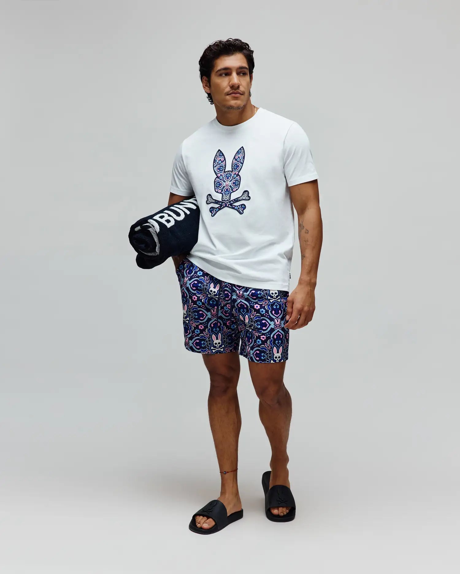 Mens Luke Graphic Tee, a classic fit white jersey t-shirt with a large, vibrant patterned Psycho Bunny skull chest graphic, paired with relaxed navy shorts featuring an all-over bunny skull and floral print.