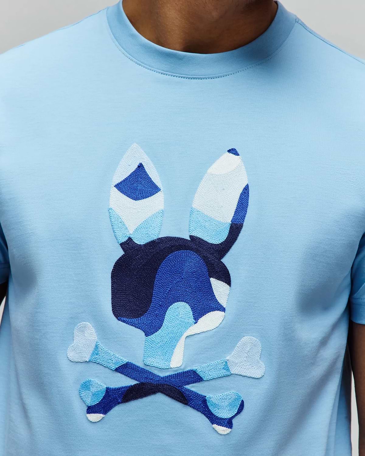 Mens Ara Graphic Tee, a light blue jersey t-shirt with a large, central embroidered Psycho Bunny skull logo in shades of blue and white camouflage, classic fit.