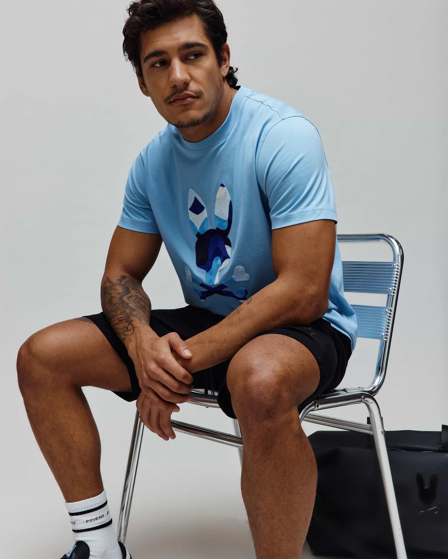 Mens Ara Graphic Tee, a light blue classic fit jersey knit t-shirt featuring a large, central graphic of the iconic Psycho Bunny skull logo in shades of blue, white, and black.